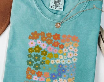 May include: A turquoise Comfort Colors t-shirt featuring a colorful floral design. The design includes flowers in shades of orange, pink, yellow, green, and blue. The text "CELEBRATE MINDS OF ALL KINDS" is printed below the flowers. A gold necklace is draped over the shirt.