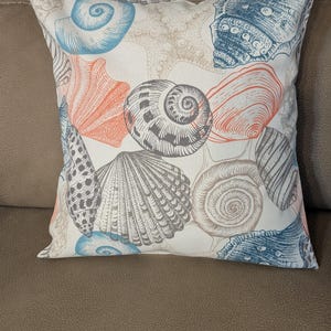 May include: Square decorative pillow with a white background and a seashell pattern. The seashells are in shades of blue, coral, gray, and tan. The pillow is resting on a brown surface.