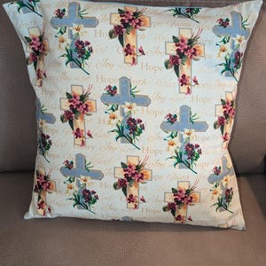 May include: A decorative pillow with a cream-colored background featuring repeating patterns of crosses, floral arrangements, and script lettering. The crosses are light blue and beige, adorned with burgundy and white flowers. The words "Faith," "Hope," and "Joy" are visible.
