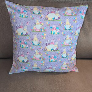 May include: Square decorative pillow with a lavender background featuring a repeating pattern of Easter gnomes and bunnies. The gnomes wear hats and hold Easter eggs, surrounded by colorful flowers. The pillow is on a brown surface.