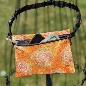 May include: An orange and yellow sunflower print crossbody bag with a black adjustable strap and zipper closure. The bag contains a smartphone, keys, and cash. The bag is hanging on a black metal display stand.