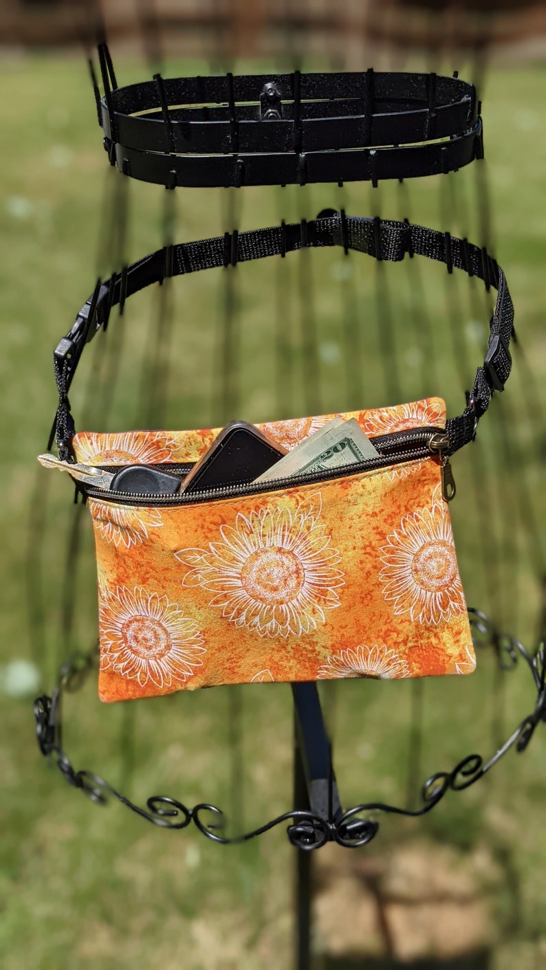 May include: An orange and yellow sunflower print crossbody bag with a black adjustable strap and zipper closure. The bag contains a smartphone, keys, and cash. The bag is hanging on a black metal display stand.
