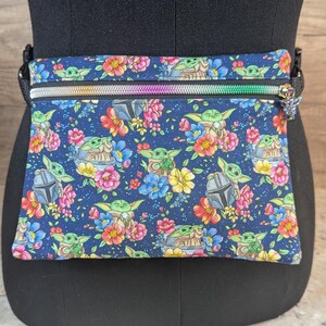May include: A navy blue fanny pack with a colorful floral and character print. The design features a repeating pattern of a green alien character, flowers, and a helmet. The pack has a rainbow zipper and a black adjustable strap.