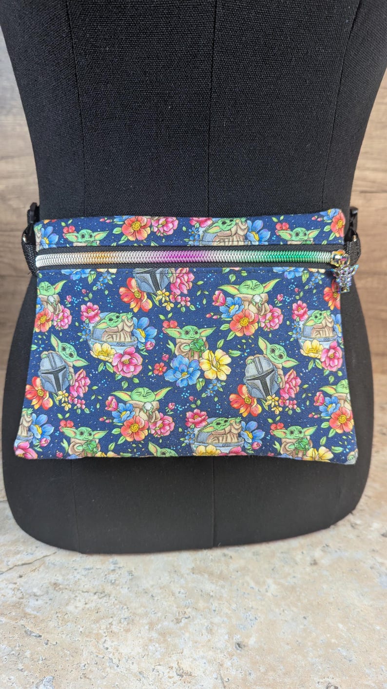 May include: A navy blue fanny pack with a colorful floral and character print. The design features a repeating pattern of a green alien character, flowers, and a helmet. The pack has a rainbow zipper and a black adjustable strap.
