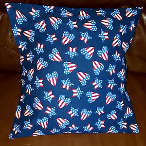 May include: Square pillow with a navy blue background, covered in a pattern of red, white, and blue stars and hearts. The pillow is a decorative item, perfect for adding a patriotic touch to any room.