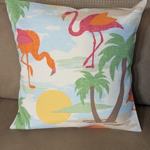 May include: Square decorative pillow with a tropical design. The pillow features two flamingos, palm trees, and a setting sun against a light blue and white background. The flamingos are pink and orange.