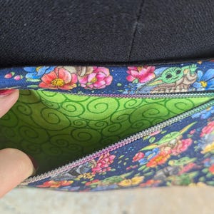 May include: A blue floral crossbody bag with a green interior. The bag features a colorful pattern of flowers and a cartoon character. The bag has a zipper closure and a black strap.
