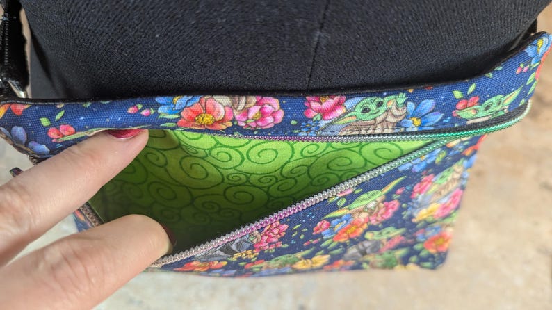 May include: A blue floral crossbody bag with a green interior. The bag features a colorful pattern of flowers and a cartoon character. The bag has a zipper closure and a black strap.
