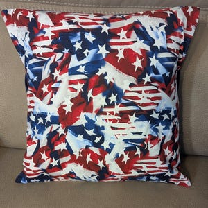 May include: Square pillow with a patriotic design featuring red and white stripes and white stars on a blue background. The pillow has a soft, textured appearance and is likely made of fabric.