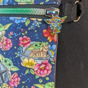 May include: A navy blue fanny pack with a colorful floral and Grogu print. The bag has a green zipper and a Grogu charm. The strap is black. The flowers are pink, red, yellow, and blue.