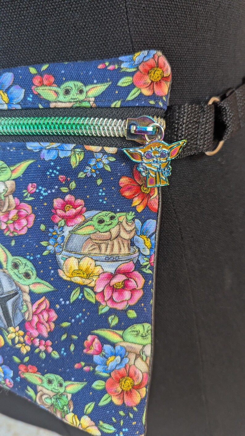 May include: A navy blue fanny pack with a colorful floral and Grogu print. The bag has a green zipper and a Grogu charm. The strap is black. The flowers are pink, red, yellow, and blue.