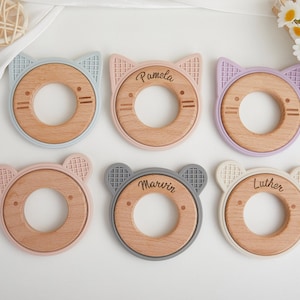 Op de afbeelding: Zes ronde bijtringen met houten centra en siliconen randen in de vorm van katten- of berenoren. Drie ringen zijn gegraveerd met de namen Pamela, Marvin en Luther. De siliconen randen zijn uitgevoerd in lichtblauw, roze, paars en grijs.