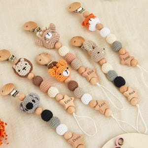 Può includere: Una collezione di clip per ciuccio per bambini con perline di legno e decorazioni a forma di animali all'uncinetto. Le clip presentano una volpe, un orso, un koala e un leone. Ogni clip ha una stella di legno con un nome inciso.