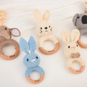 May include: A collection of handmade crochet baby rattles in the shape of animals, including an elephant, koala, and rabbits. Each rattle features a wooden ring and is in neutral colors. The names Alea, Clark, Zayn, and Dear are engraved on the wooden rings.