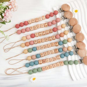 Puede incluir: Una colección de clips de chupete personalizados para bebés en varios colores, incluyendo rojo, rosa, azul y verde. Cada clip presenta cuentas de madera y un clip de madera, con nombres escritos en bloques de letras de madera. Los nombres incluyen ENOCH, BELLA, SARAH, THOMAS, CHASEL, KELLY, ANTONY y HARLEY.