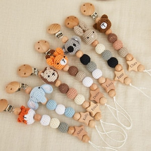 May include: A collection of handmade baby pacifier clips. Each clip features a wooden star with a name engraved on it, and a variety of crochet animal designs, including a fox, bear, koala, and elephant. The clips are in various colors.