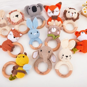 May include: A collection of handmade crochet baby rattles in various animal shapes, including a bunny, lion, fox, koala, bear, elephant, and bee. Each rattle features a wooden ring and is crafted in soft, neutral colors.