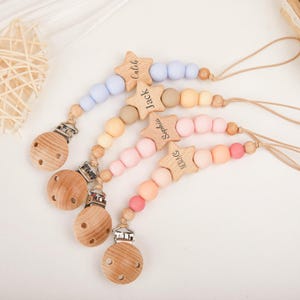 May include: Four baby pacifier clips with wooden star details, each with a different name: Caleb, Jack, Sophia, and Nora. The clips have pastel-coloured silicone beads and wooden pacifier holders. The clips are designed for babies.