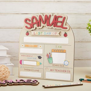 May include: A light-coloured wooden arch-shaped sign with the name SAMUEL in red letters at the top. The sign features text including LAST DAY OF, DATE, I AM, YEARS OLD, WHEN I GROW UP I WANT TO BE, THINGS I LOVE, and MY TEACHER IS with blank spaces.