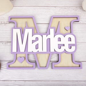 May include: A wooden sign featuring the name "Marlee" in white, layered over a lavender-colored letter "M". The letter "M" has a natural wood grain background and includes two small heart cutouts. A decorative item for a nursery or bedroom.