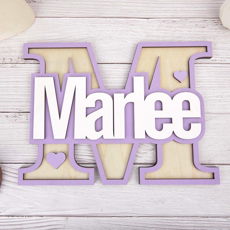 May include: A wooden sign featuring the name "Marlee" in white, layered over a lavender-colored letter "M". The letter "M" has a natural wood grain background and includes two small heart cutouts. A decorative item for a nursery or bedroom.