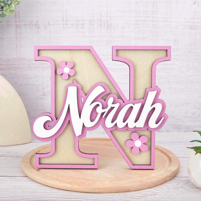May include: A wooden letter "N" with the name "Norah" in white script, accented with pink trim and small flower details. The wooden cutout is light beige with pink trim and sits on a wooden tray.