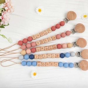 May include: Four personalized baby pacifier clips with wooden beads and names spelled out in wooden letter blocks. The clips feature a variety of colors, including pinks, blues, and neutrals, with wooden clip ends.