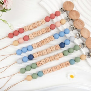 May include: A collection of personalized baby pacifier clips in various colors. Each clip features wooden beads, silicone beads, and a wooden clip. Names are spelled out with wooden letter blocks. Colors include red, pink, blue, green, and tan.