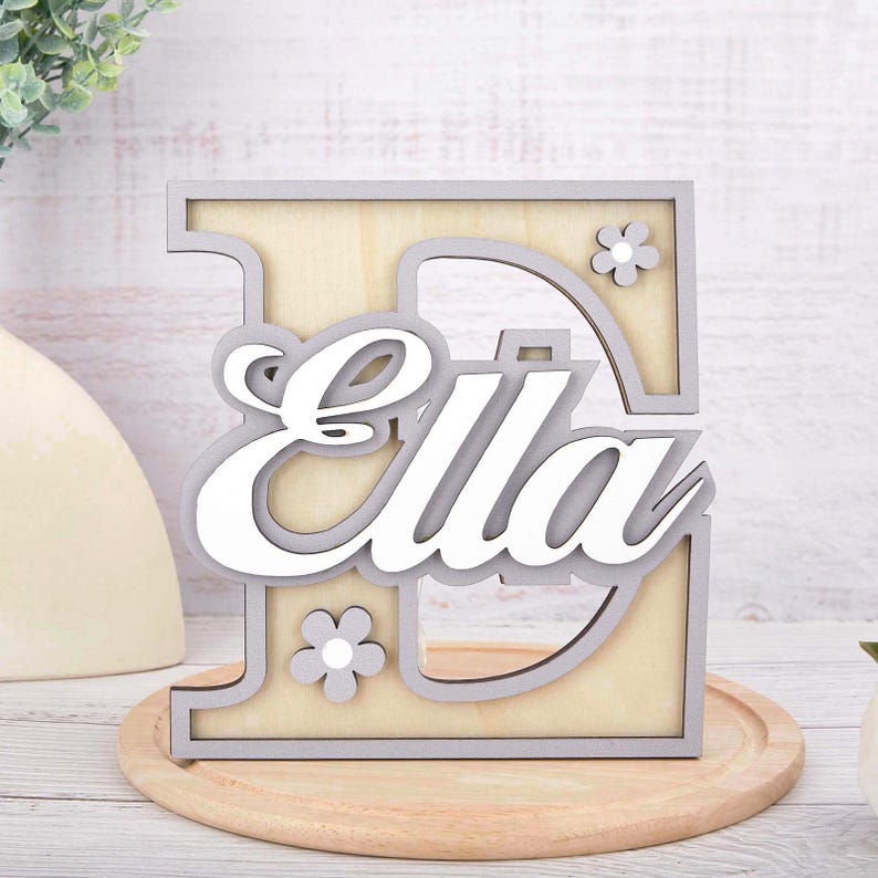May include: Wooden letter "E" with the name "Ella" in white script. The letter is outlined in gray, with a light wood interior and two small flower accents. The piece is displayed on a wooden tray.
