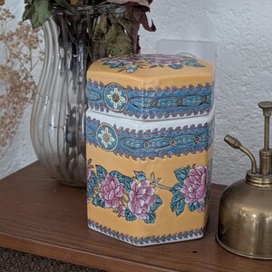 Puede incluir: Un tarro de cerámica hexagonal con tapa, decorado con motivos florales rosas, azules y verdes sobre fondo amarillo. Un pulverizador de latón vintage está al lado del tarro. Ideal para el almacenamiento.