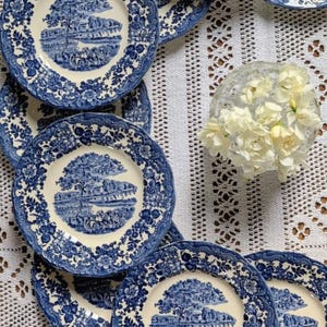 Pode incluir: Um conjunto de pratos de porcelana azuis e brancos com um design paisagístico. Os pratos apresentam uma imagem central de uma paisagem rodeada por uma borda floral. Estão dispostos sobre uma toalha de renda branca, com um pequeno vaso de flores brancas.
