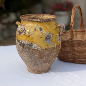 Può includere: Un vaso in ceramica bulboso e usurato, caratterizzato da una sezione superiore smaltata in giallo vivace e una base strutturata color marrone terra. Il recipiente presenta due piccole maniglie laterali, un bordo circolare e segni d'uso.