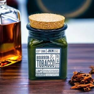 May include: A green glass jar candle with a cork lid. The label reads "EST 2008 JKM SOY candles AMPERSAND COLLECTION BOURBON & TOBACCO 100% SOY 8.5 OZ No. 6 CRACKLING ROUND WOODEN WICKS HAND-POURED IN SMALL BATCHES".
