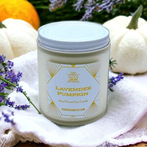 May include: A white frosted glass candle jar with a white lid. The label is gold and white with the text "Lavender Pumpkin" and "Hand Poured Soy Candle". The candle is sitting on a white towel with a white pumpkin and a bunch of lavender flowers.