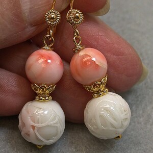 May include: A pair of dangle earrings featuring peach and white beads. The top beads are peach with white marbling, while the bottom beads are white with carved floral designs. Gold-colored accents and ear wires complete the design.