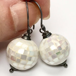 May include: A pair of white mosaic ball earrings with dark gray metal hooks and accents. The earrings are round and made of small, square tiles that reflect light. The hooks are curved and simple.