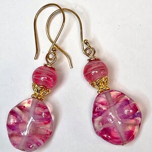 May include: A pair of handmade earrings featuring gold-colored ear wires. Each earring has a small, round, pink and white striped bead above a larger, irregular-shaped bead with pink and purple swirls. Gold-colored filigree accents are between the beads.