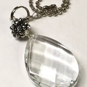 May include: A clear, faceted, teardrop-shaped glass pendant attached to a silver-toned metal ball chain. The pendant is connected to the chain by a silver wire-wrapped cluster of small, dark gray, faceted beads and a circular metal jump ring.