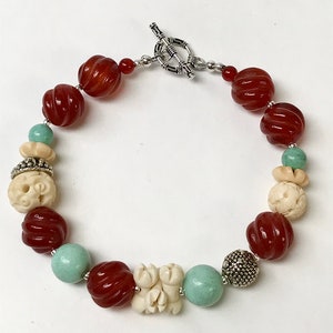 May include: A red, white, and green beaded necklace with a silver toggle clasp. The necklace features carved bone beads, red carnelian beads, and green jade beads.