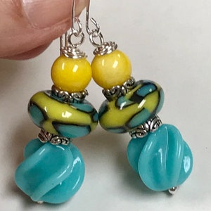 May include: A pair of handmade earrings featuring a combination of yellow, teal, and silver beads. The earrings have a unique design with a mix of round and textured beads, creating a vibrant and eye-catching accessory.