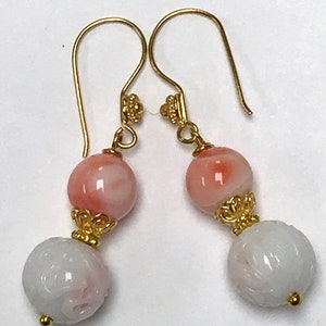 May include: A pair of drop earrings featuring gold-tone hooks and decorative caps. Each earring consists of a smooth, round, peach-colored bead suspended above a larger, white, spherical bead with an intricate, carved floral pattern.
