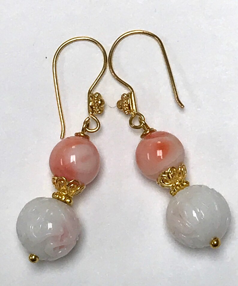 May include: A pair of drop earrings featuring gold-tone hooks and decorative caps. Each earring consists of a smooth, round, peach-colored bead suspended above a larger, white, spherical bead with an intricate, carved floral pattern.