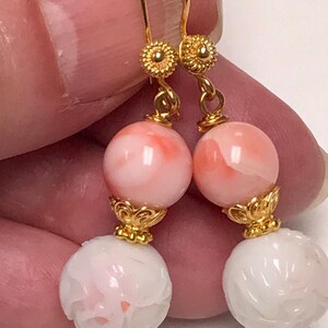 May include: A pair of drop earrings featuring gold-tone hooks with textured circular accents. Each earring displays a smooth, round, pale pink bead above a white, spherical bead with an intricate carved floral pattern, connected by gold-tone findings.