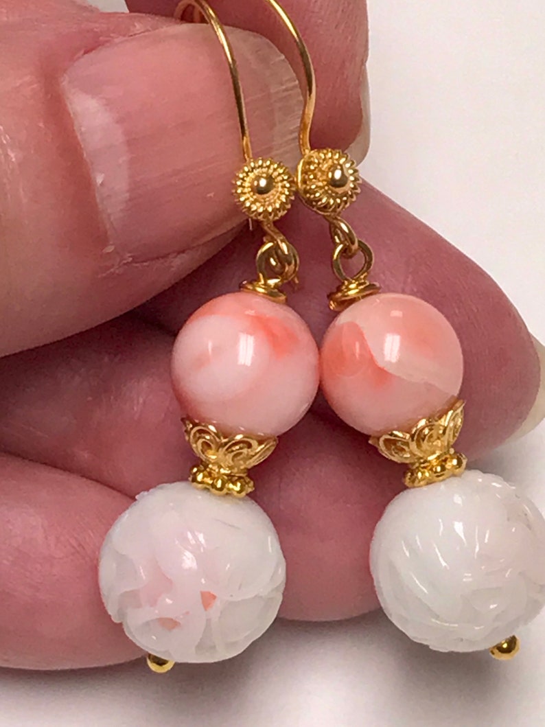 May include: A pair of drop earrings featuring gold-tone hooks with textured circular accents. Each earring displays a smooth, round, pale pink bead above a white, spherical bead with an intricate carved floral pattern, connected by gold-tone findings.