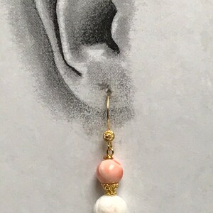 May include: A gold-toned dangle earring featuring a smooth, round, light pink bead positioned above a textured, round, white bead. The beads are separated and accented by small, intricate gold-toned metal caps, suspended from a gold-toned hook wire.
