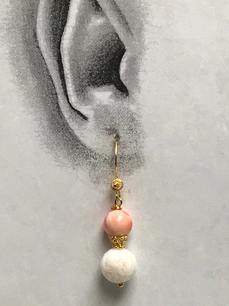 May include: A gold-toned dangle earring featuring a smooth, round, light pink bead positioned above a textured, round, white bead. The beads are separated and accented by small, intricate gold-toned metal caps, suspended from a gold-toned hook wire.