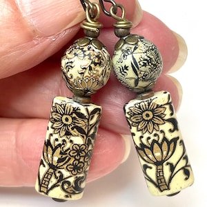 May include: A pair of drop earrings featuring cylindrical and spherical beads in cream with black and gold floral patterns. The beads are accented with bronze-toned metal caps and wire wraps, suspended from dark metal ear wires for a dangling effect.