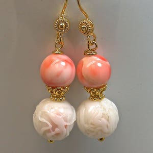 May include: A pair of drop earrings featuring gold-tone ear wires with floral accents. Each earring consists of a smooth, round, light pink bead suspended above a larger, white, spherical bead with intricate, carved, raised floral patterns.