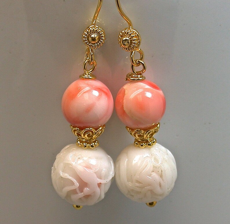 May include: A pair of drop earrings featuring gold-tone ear wires with floral accents. Each earring consists of a smooth, round, light pink bead suspended above a larger, white, spherical bead with intricate, carved, raised floral patterns.