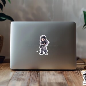 Puede incluir: Una pegatina con una ilustraci&oacute;n de dibujos animados de una persona joven que lleva un atuendo blanco y negro y un sombrero negro. La pegatina est&aacute; en un ordenador port&aacute;til.